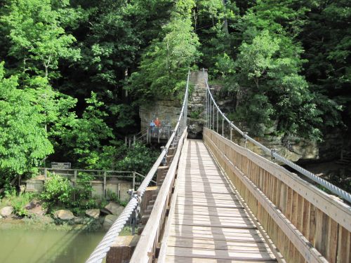 A wooden suspension bridge spans a river, surrounded by lush green trees and rocky cliffs.