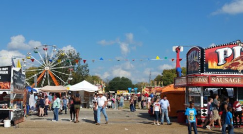 A lively fair scene with food stalls, a Ferris wheel, and crowds enjoying the festivities under a blue sky.