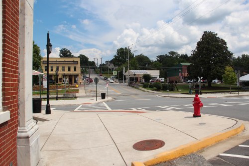 A quiet town intersection with a fire hydrant, buildings, and trees under a partly cloudy sky.