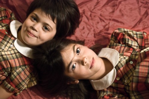 Two girls in matching plaid dresses lie on a pink surface, smiling and looking at the camera.