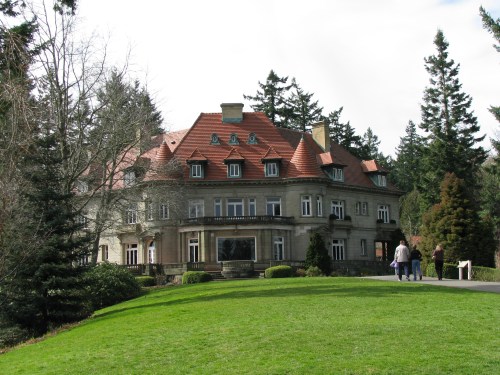 A large, elegant mansion with a red-tiled roof, surrounded by trees and a green lawn, with people walking nearby.