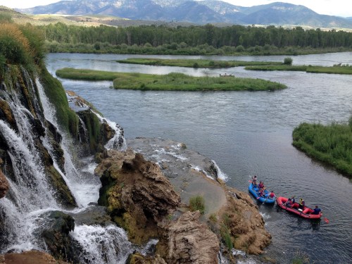 A scenic view of a waterfall cascading into a river, with a group in rafts nearby and mountains in the background.