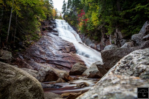 A serene waterfall cascades over rocky terrain, surrounded by vibrant autumn foliage and smooth flowing water.