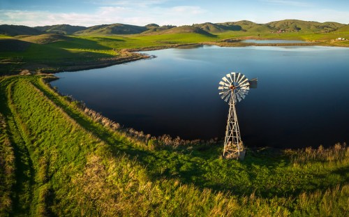 A serene landscape featuring a windmill by a calm lake, surrounded by lush green hills under a clear blue sky.