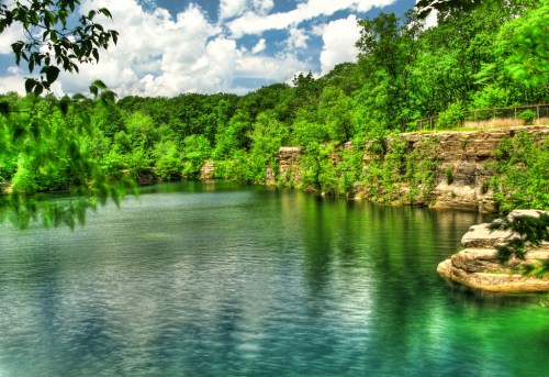 A serene lake surrounded by lush greenery and rocky cliffs under a bright blue sky with fluffy clouds.