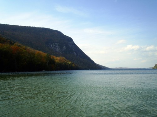A serene lake surrounded by mountains, with clear blue water and a partly cloudy sky. Autumn colors on the trees.