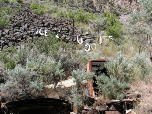 Abandoned structures and rusted metal amidst overgrown vegetation and rocky terrain.