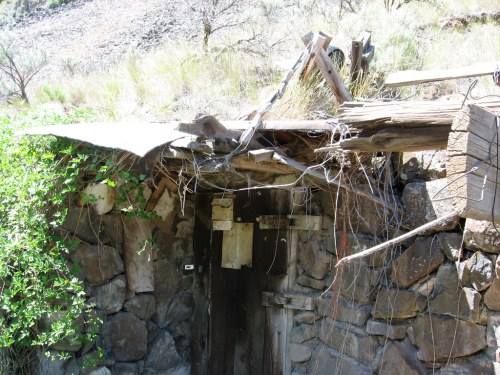 An old, weathered stone structure with a partially collapsed roof, surrounded by dry vegetation and rocks.