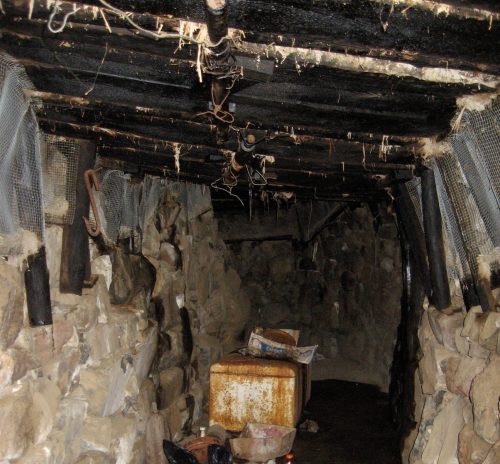 A dimly lit, narrow underground tunnel with stone walls, debris, and scattered trash on the floor.