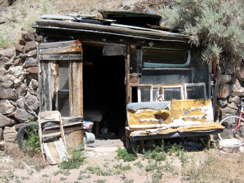 A dilapidated wooden shack with a worn-out bench outside, surrounded by rocky terrain and sparse vegetation.
