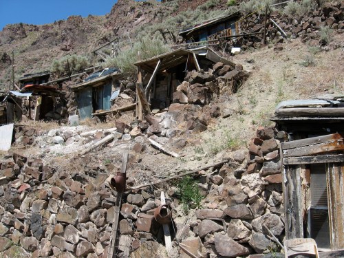 Abandoned stone structures on a hillside, surrounded by rocky terrain and sparse vegetation under a clear blue sky.