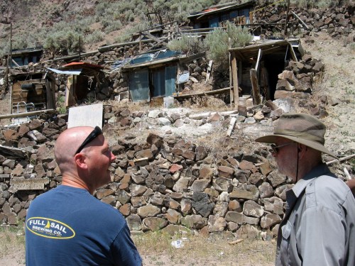 Two men stand in front of a rocky hillside with dilapidated buildings and vegetation in the background.