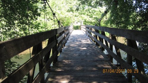 A wooden suspension bridge surrounded by lush greenery, leading over a calm waterway.