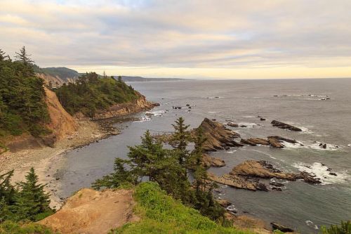 A scenic coastal view with rocky shores, lush greenery, and a calm ocean under a cloudy sky.