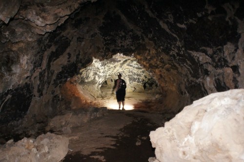 A person stands in a dark cave, illuminated by a flashlight, surrounded by rocky walls and a sandy floor.