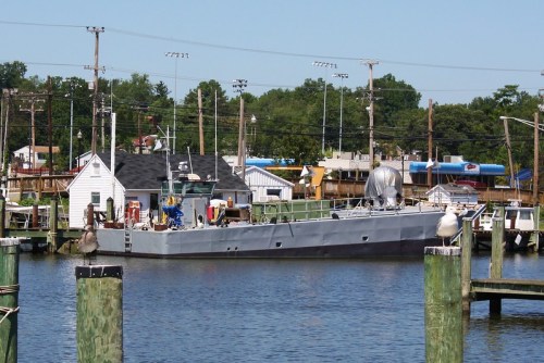 A gray boat docked at a marina, surrounded by water and buildings, with seagulls perched on wooden posts.
