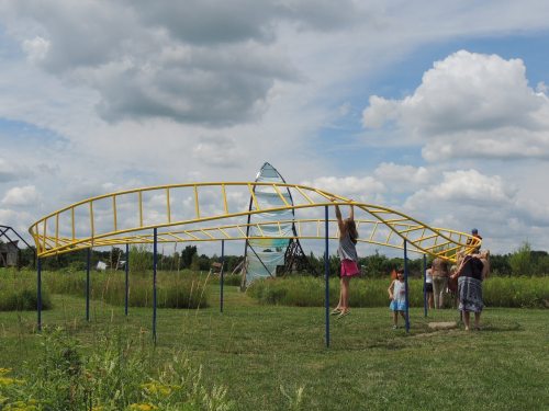 Children playing on a yellow playground structure in a grassy field under a cloudy sky.