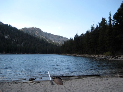 A serene lake surrounded by mountains and pine trees, with a sandy shore in the foreground.