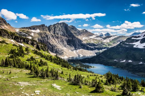 A scenic mountain landscape with lush green valleys, rocky peaks, and a serene blue lake under a bright blue sky.