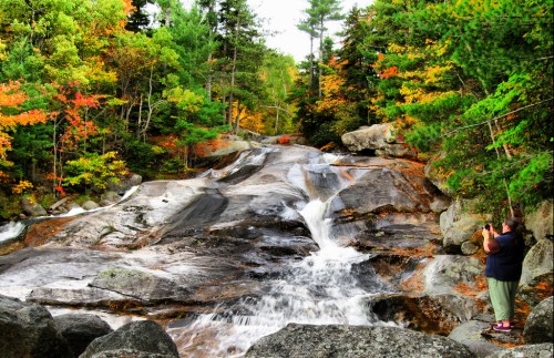 A person stands by a rocky waterfall surrounded by vibrant autumn foliage and tall trees.