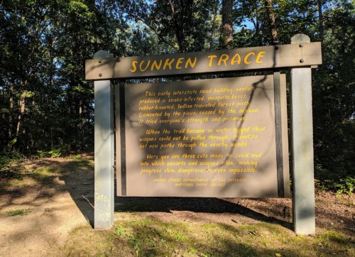 Sign for "Sunken Trace" detailing the history of an early interstate road and its challenges, surrounded by trees.