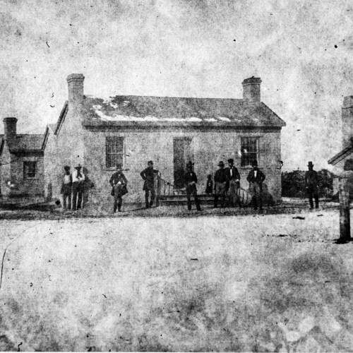 Black and white photo of a snowy scene with a group of men standing in front of a small house.