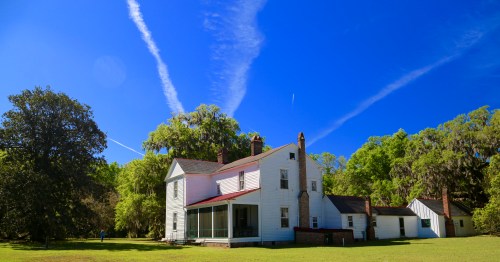 A white two-story house surrounded by green trees and a clear blue sky with wispy clouds.