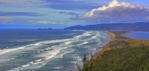 A scenic coastal view featuring waves, sandy beaches, and distant hills under a partly cloudy sky.