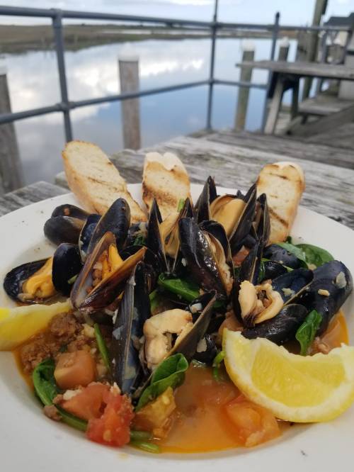 A plate of mussels in a savory broth, garnished with lemon and served with toasted bread, by a scenic waterfront.