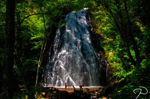 A waterfall cascades down rocky cliffs, surrounded by lush green trees, with two people standing at the base.