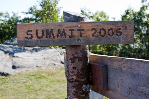 Wooden signpost reading "SUMMIT 2006'" at a scenic outdoor location, surrounded by trees and clear blue sky.