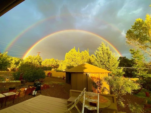A vibrant double rainbow arcs over a lush backyard with trees and a small shed, under a cloudy sky.