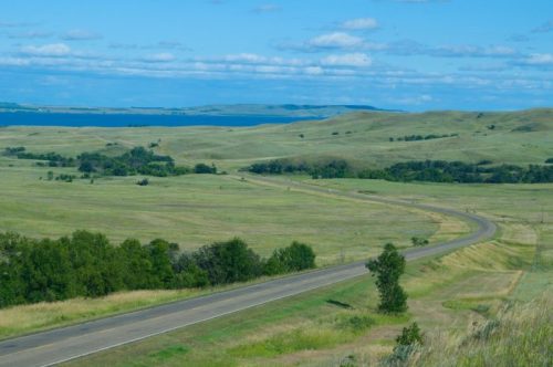 A winding road through green hills, with a lake and blue sky in the background.