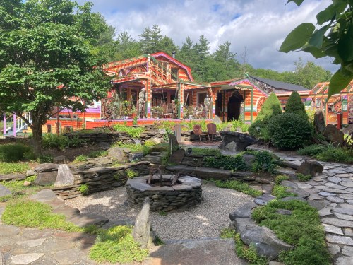 Colorful buildings surrounded by lush greenery and a stone pathway, featuring a central stone fire pit and garden area.