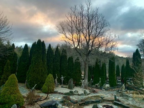 A serene garden with tall evergreen trees, a bare tree, and a stone pathway under a cloudy sky at sunset.