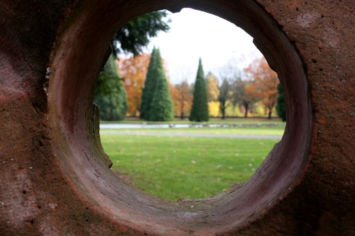View through a circular stone opening, framing a park with evergreen trees and autumn foliage in the background.