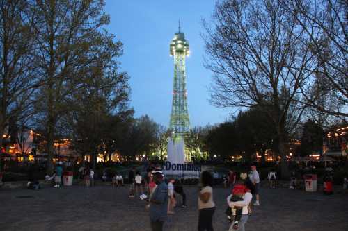 A tall illuminated tower stands in a park at dusk, surrounded by trees and people enjoying the evening.