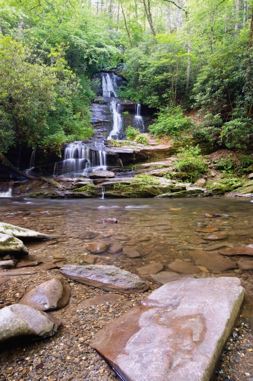 A serene waterfall cascades over rocks into a clear pool, surrounded by lush green trees and foliage.
