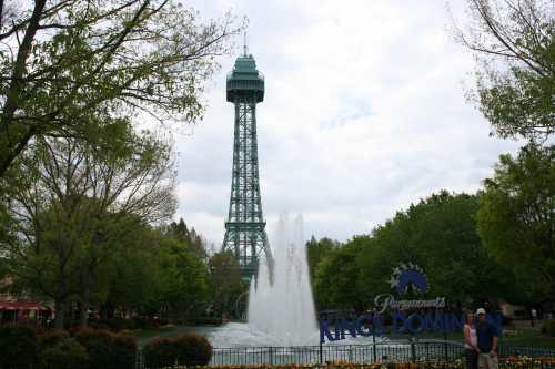 A tall amusement park tower stands behind a fountain, surrounded by lush greenery and colorful flowers.