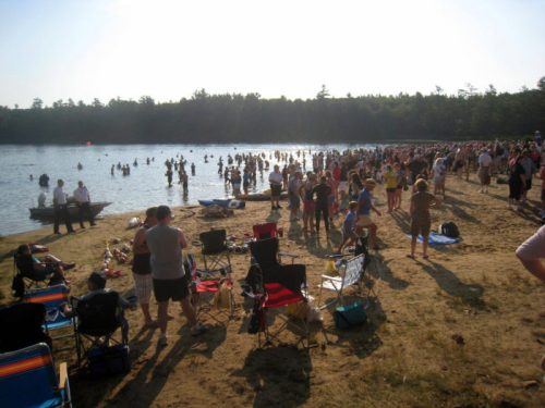A crowded beach scene with people swimming in the water and relaxing on the sand, surrounded by trees.