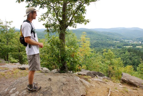 A hiker stands on a rock, overlooking a lush green valley and distant mountains under a cloudy sky.