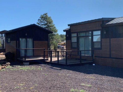 Two modern tiny homes with wooden exteriors, surrounded by gravel and trees, under a clear blue sky.