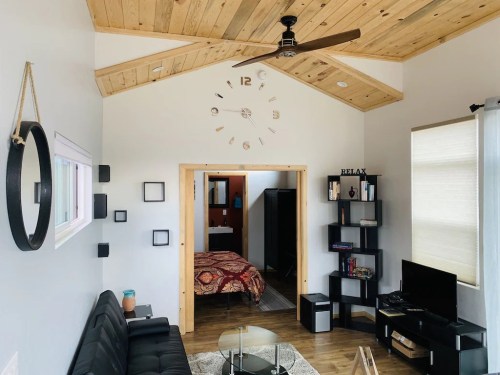 Cozy living room with wooden ceiling, clock, black sofa, TV, and a view of a bedroom through an open doorway.