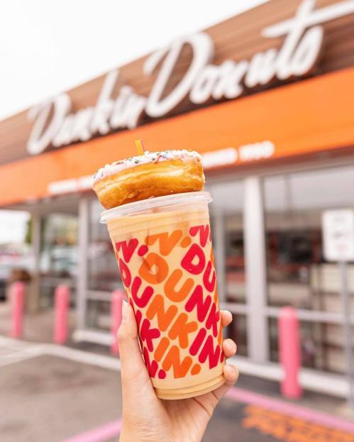 A hand holding a colorful iced drink topped with a donut, in front of a Dunkin' Donuts store.