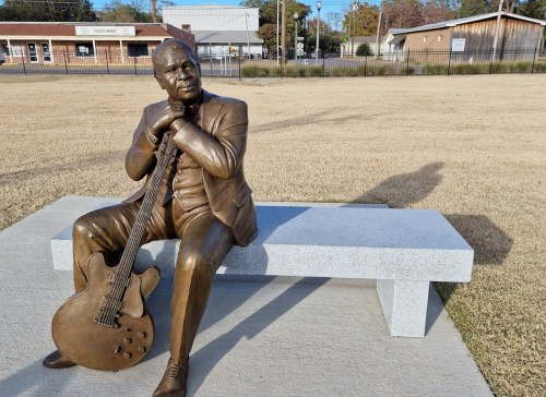 A bronze statue of a seated musician holding a guitar, placed on a stone bench in a grassy area.