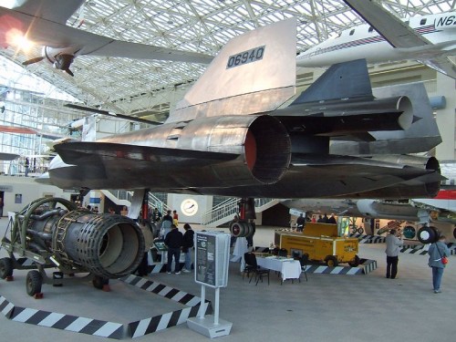 A sleek aircraft displayed indoors, showcasing its jet engine and modern design, with visitors observing nearby.