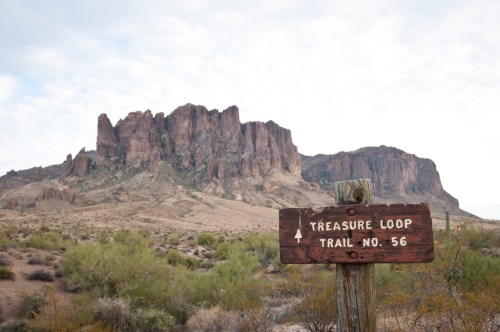 A wooden sign for Treasure Loop Trail No. 56 in front of rugged mountains and desert landscape under a cloudy sky.