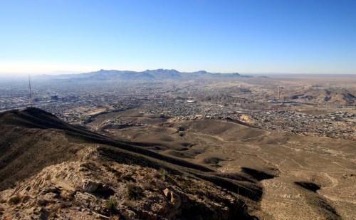 A panoramic view of a desert landscape with mountains and a city in the distance under a clear blue sky.