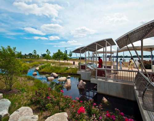 A scenic park with a walkway, stream, and blooming flowers under a blue sky with fluffy clouds.