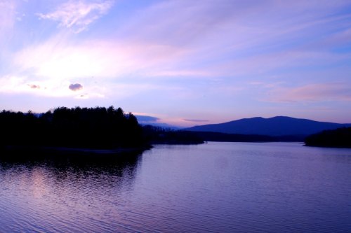A serene lake at sunset, with soft purple and blue hues reflecting on the water and mountains in the background.